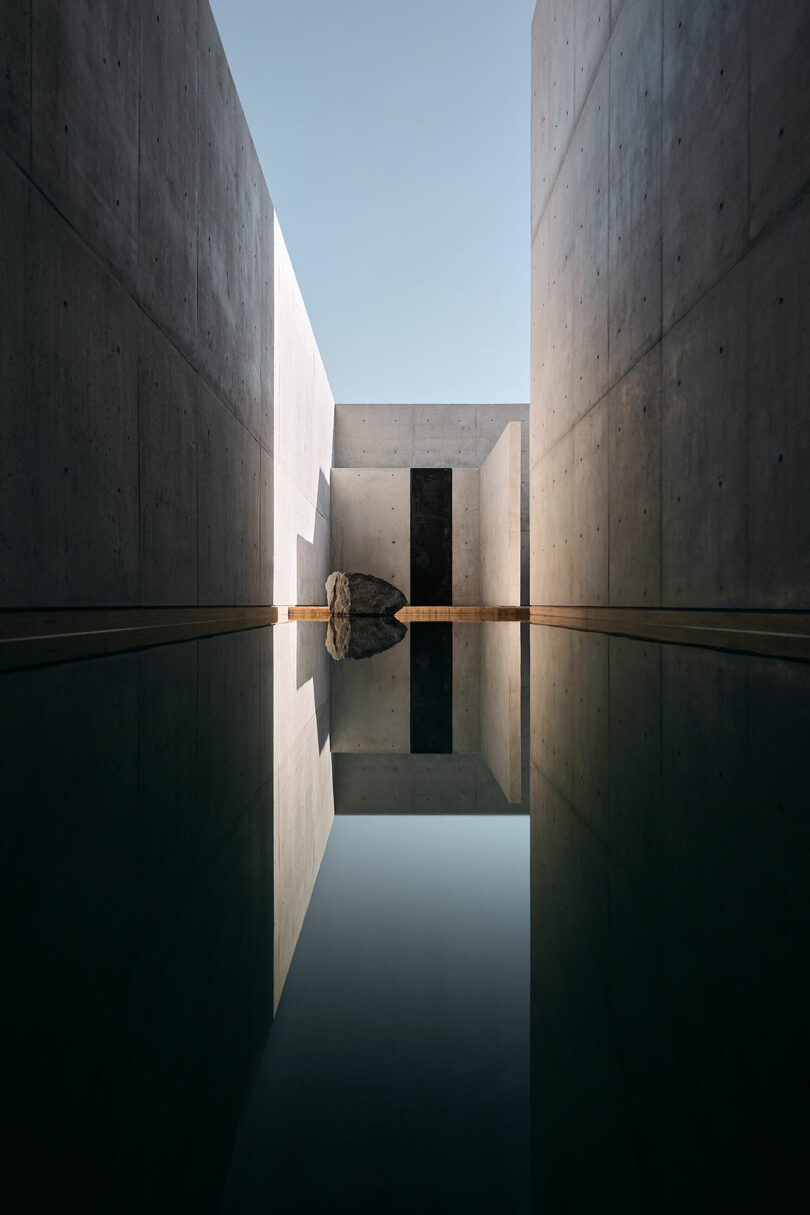 A narrow reflective pool between concrete walls with a large rock centerpiece.