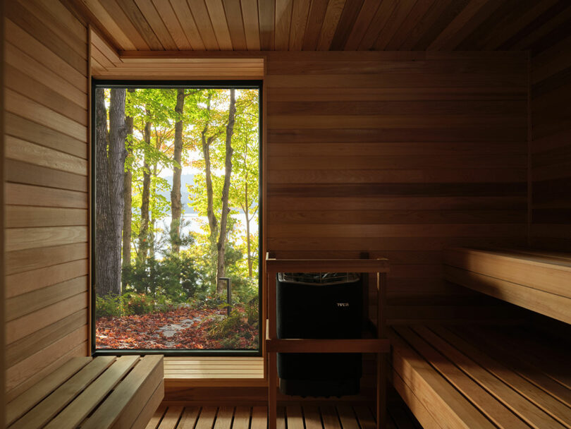 Interior of a light-wood sauna with a large window looking out onto a forest and lake.