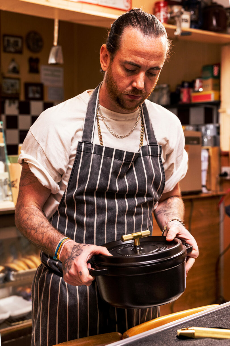 A man in a striped apron holding a Staub pot in a kitchen environment.