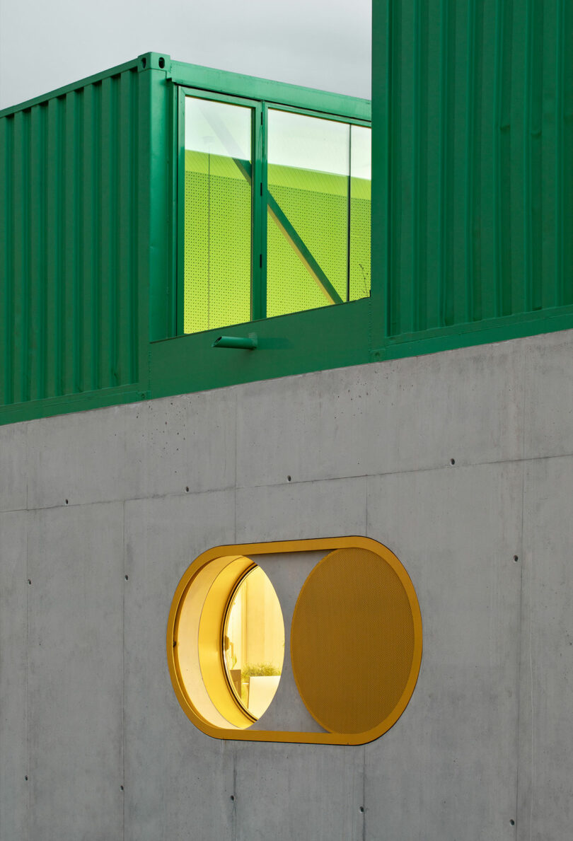 A green shipping container structure positioned atop a sturdy concrete plinth with a yellow-framed circular window.