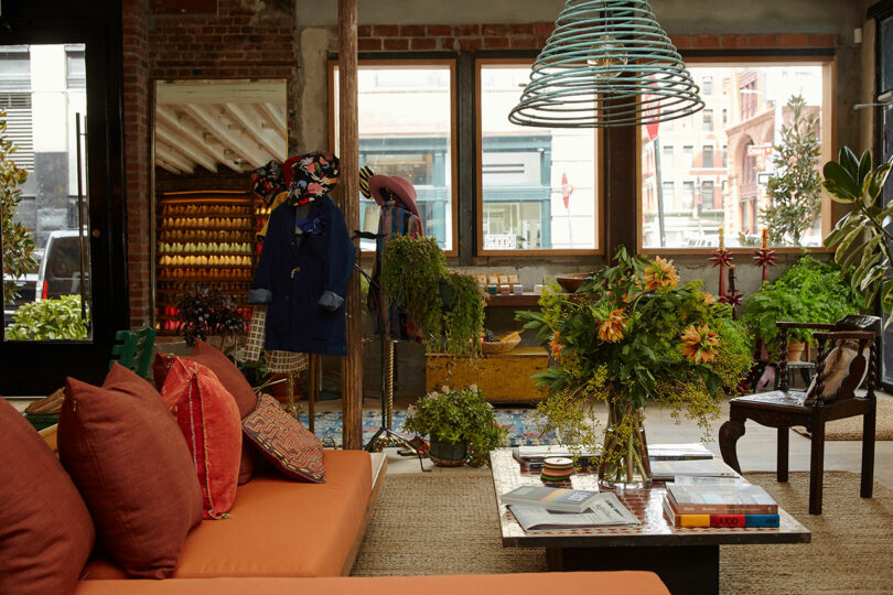 A sunlit, eclectic lounge with orange sofas, leafy plants, and a clothing display.