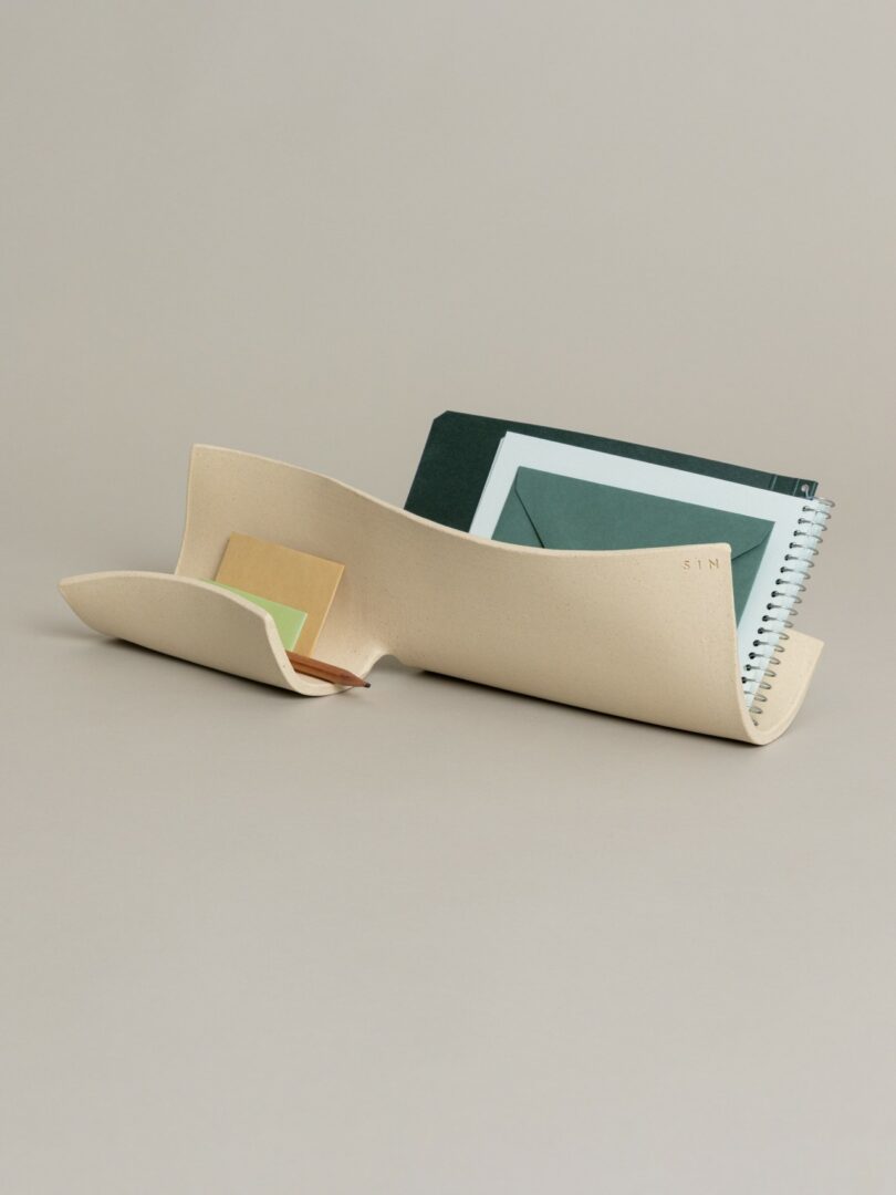 A wavy, cream-colored ceramic organizer holding a notebook and a green envelope.