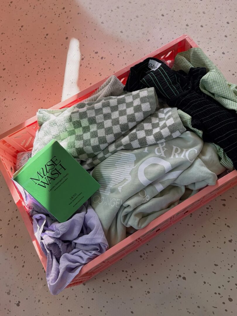 A box of MUSEWASH detergent sheets sitting on a pink crate filled with laundry.
