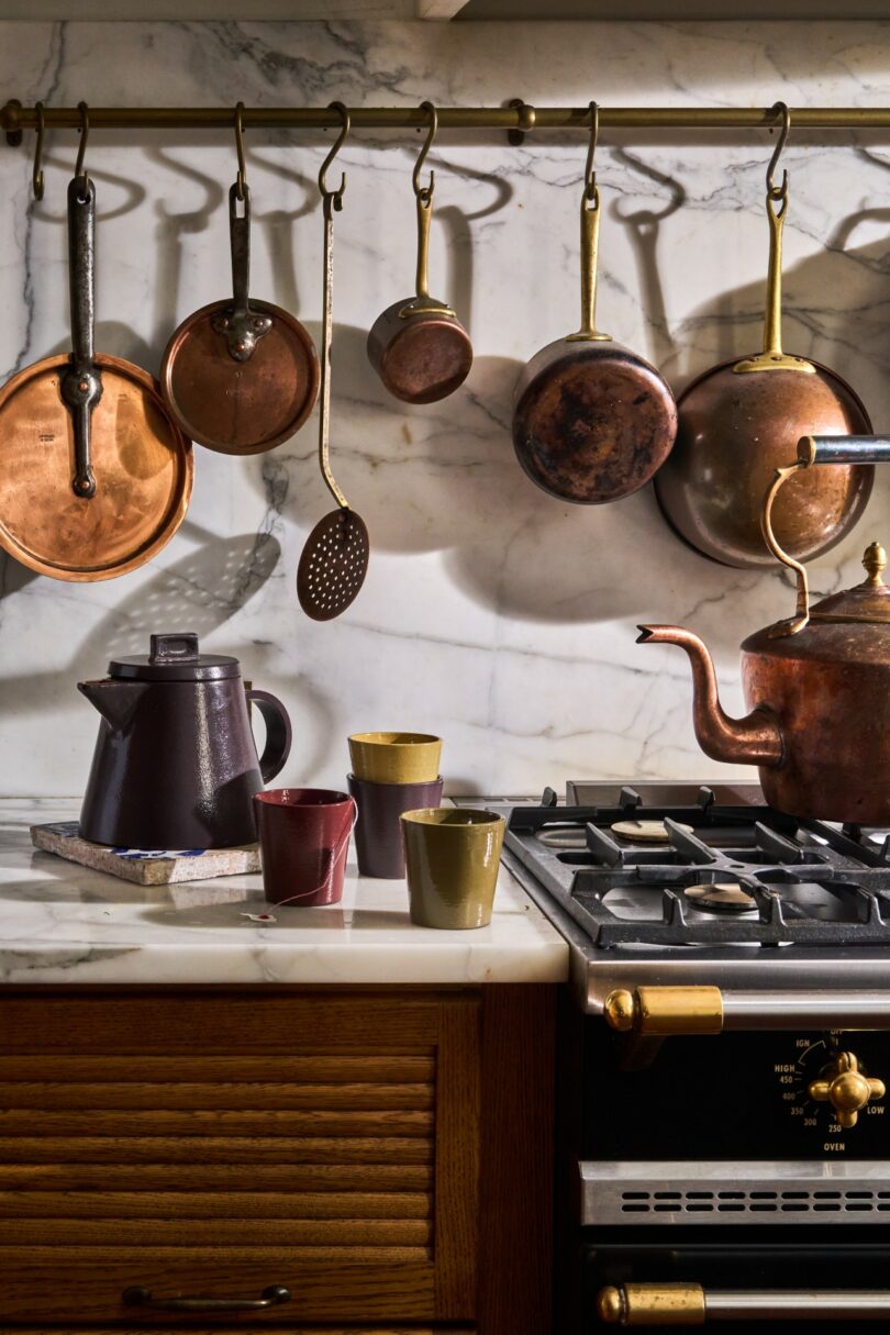 Copper pots and pans hanging above a marble countertop with ceramic mugs and a coffee pot.