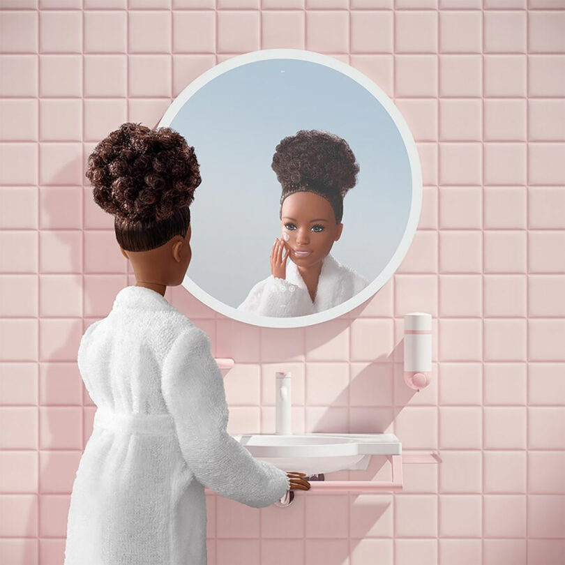 A Barbie doll in a white robe at a pink-tiled bathroom vanity with a round mirror.