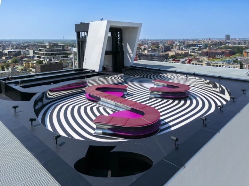 An S-shaped magenta bench on a striped floor overlooking the urban skyline of Milan under a blue sky.