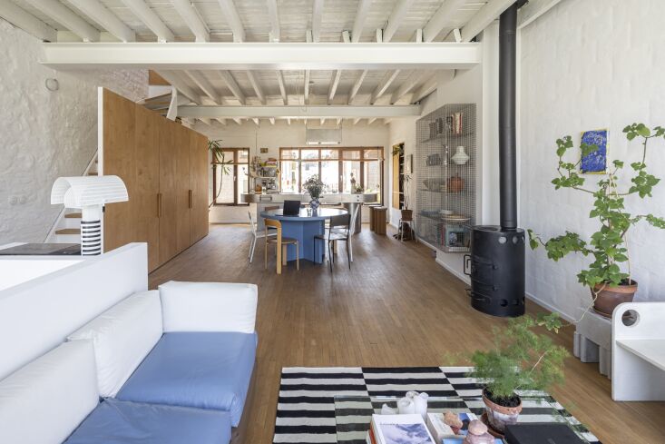 An open-plan living room featuring a blue designer sofa, steel beams, and birch plywood partitions.