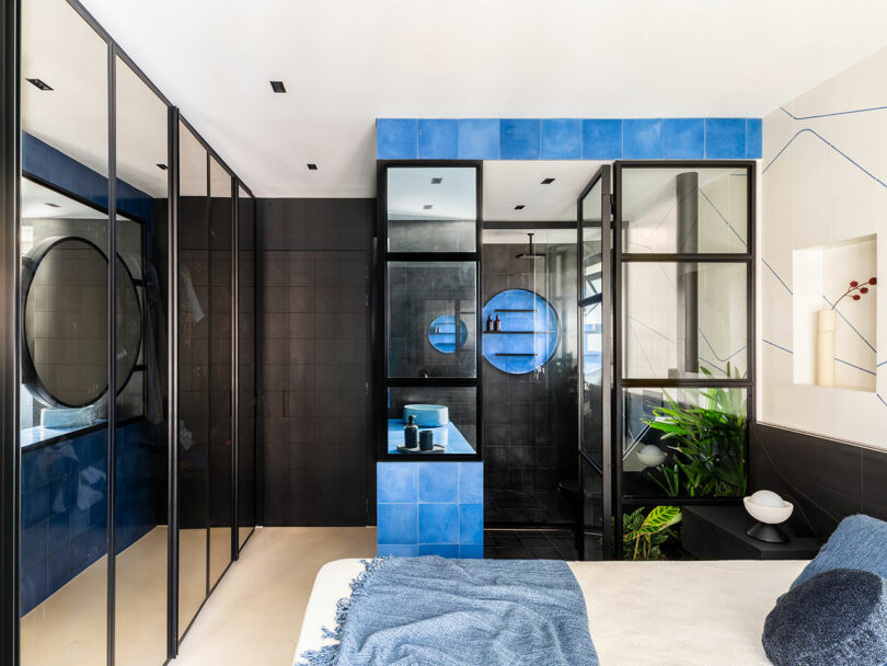 Modern bathroom with blue tiles and glass partitions adjacent to a bed with a blue throw.