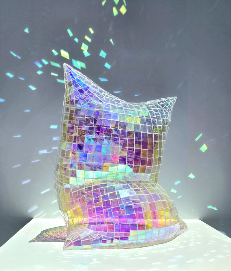 A sculpted iridescent pillow covered in mosaic tiles reflecting a spectrum of colors on a white surface.