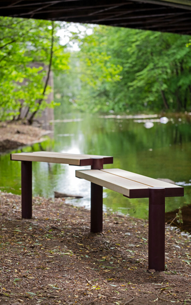 Two wooden modular benches placed along a riverbank with a bridge in the background.
