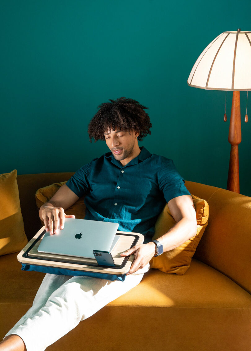 A man sitting on a sofa struggling to balance a laptop, iPad, and iPhone on his lap without a desk.