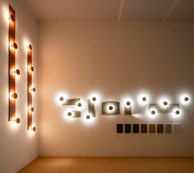 Multiple geometric wall light configurations mounted on a beige wall above a row of color finish samples.