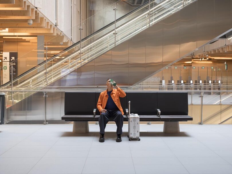 A man sitting with a suitcase in a modern transit hub, drinking from an Elio bottle.