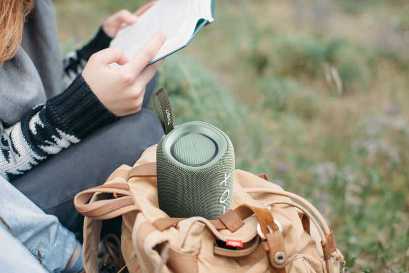 A green TRIBIT speaker resting on a backpack outdoors next to a book.