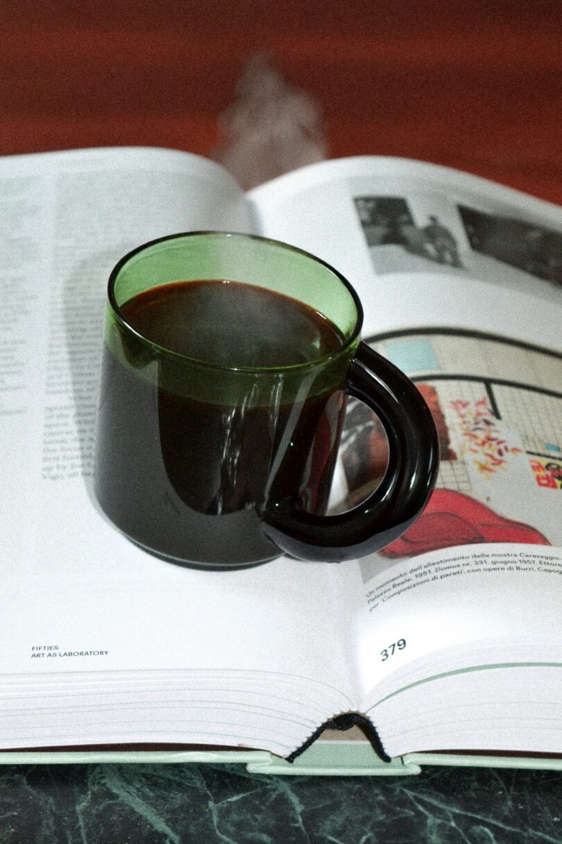 Steam rises from a green-rimmed glass mug filled with black coffee resting on an open book.