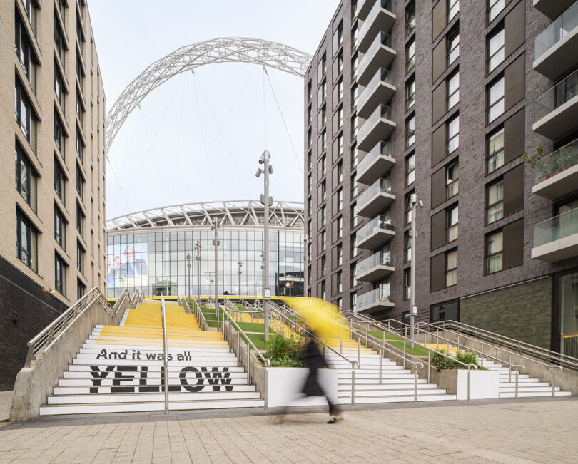 A person with a yellow umbrella walks up the steps with the Wembley Stadium arch framed by modern buildings in the background.