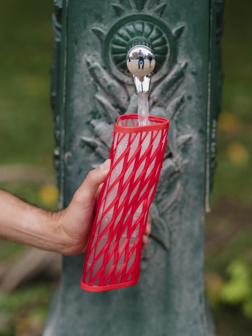 A hand holding a red Elio water bottle under a public water fountain spout.