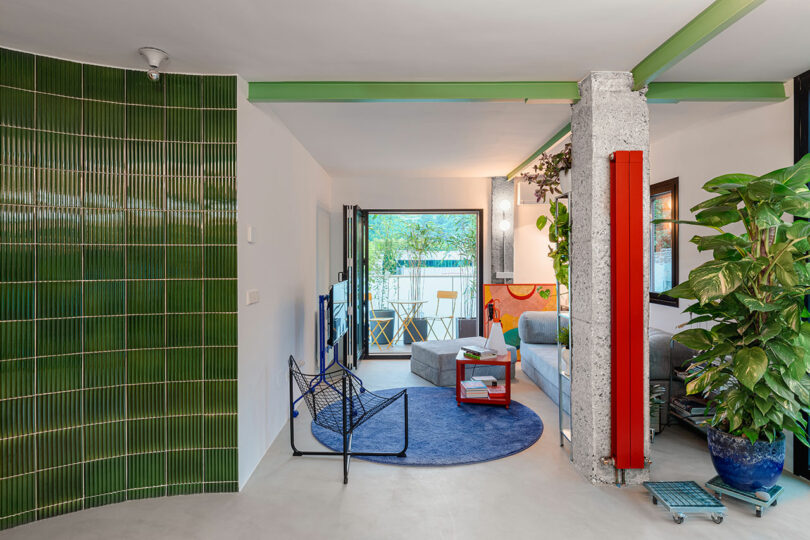 An open-plan living room with green tiled walls, a blue rug, and large windows allowing natural light to enter.
