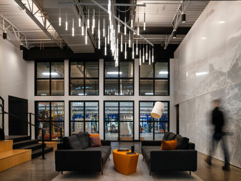 A modern lobby with dark sofas where a person walks by a large window looking into the industrial workshop.