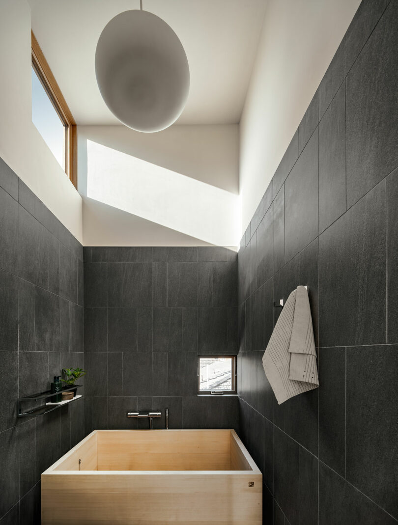 A minimalist bathroom featuring a deep light-wood soaking tub against dark tiled walls.