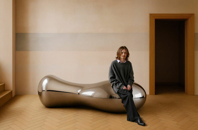 A person sitting on a shiny, bean-shaped metallic bench in a minimalist room.