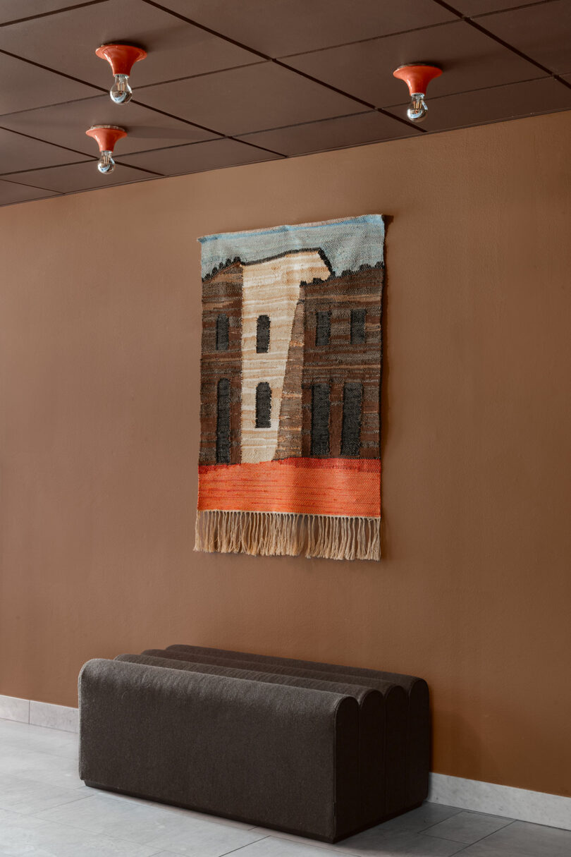 A woven wall tapestry depicting a building hanging above a dark upholstered bench with red ceiling lights.