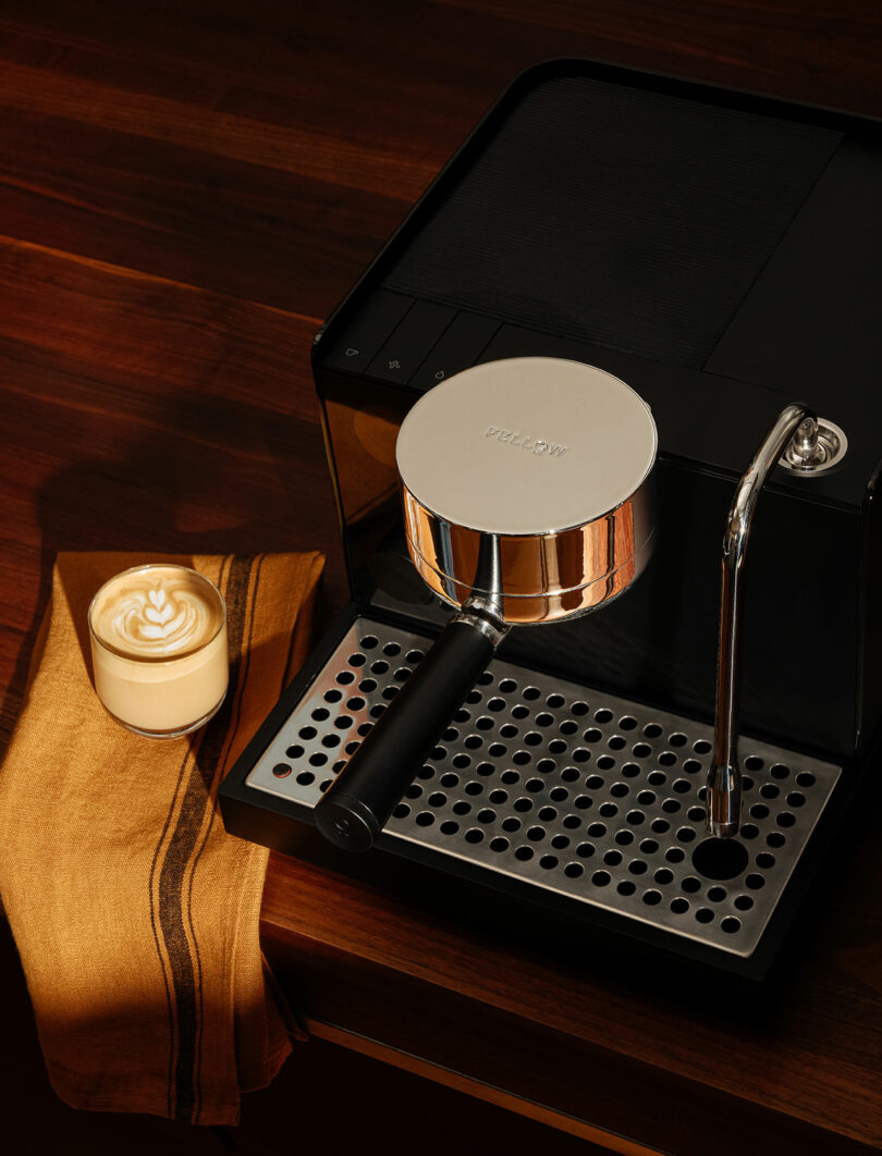 A black Fellow espresso machine on a wooden counter next to a glass of latte with latte art.