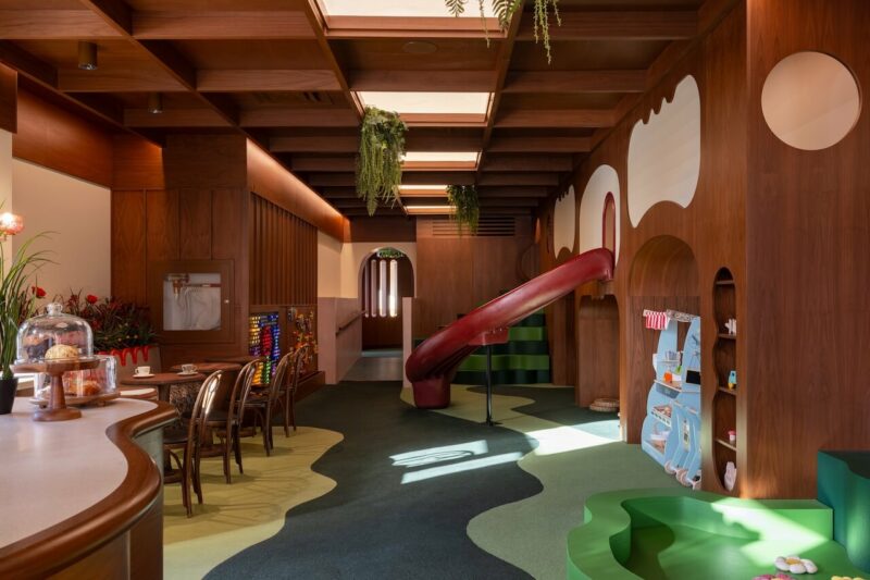 Indoor children's playroom with a bright red slide, wooden climbing structures, and hanging greenery.