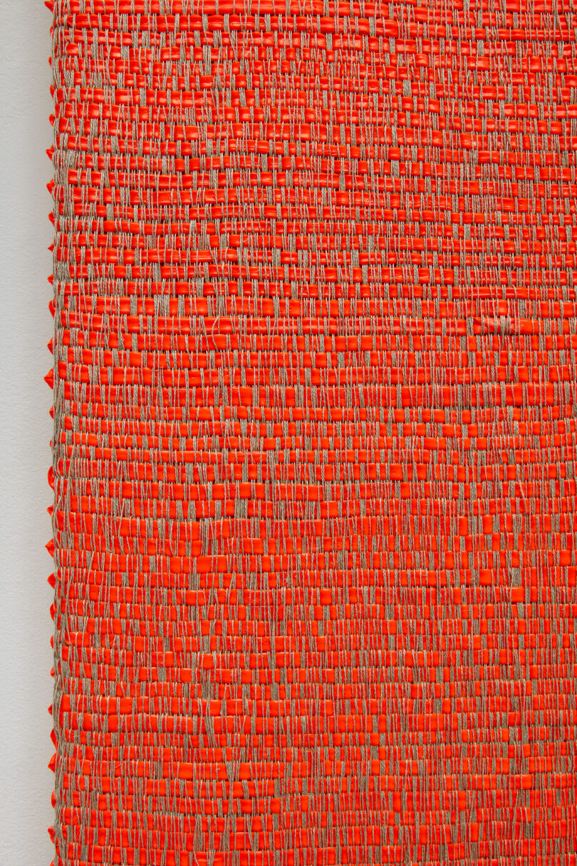 An extreme close-up showing thick strands of fluorescent orange acrylic paint interlaced in a basket-weave pattern.