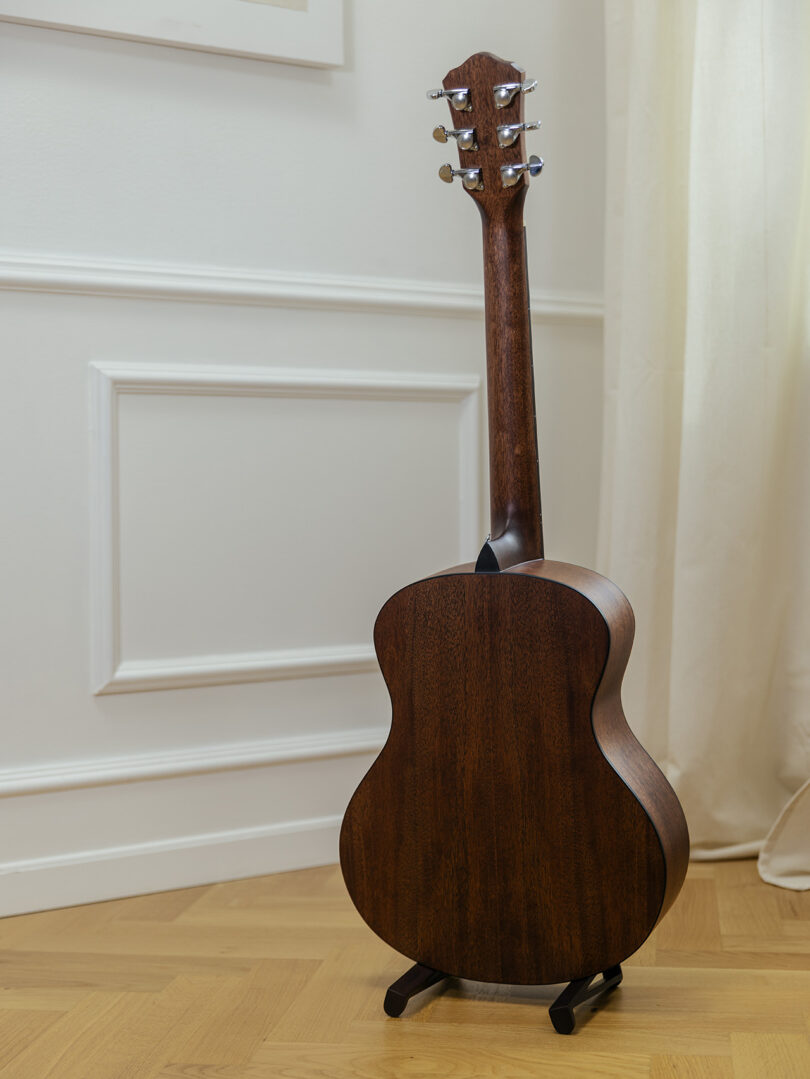 Rear view of a wooden acoustic guitar on a black minimalist stand against a cream wall.