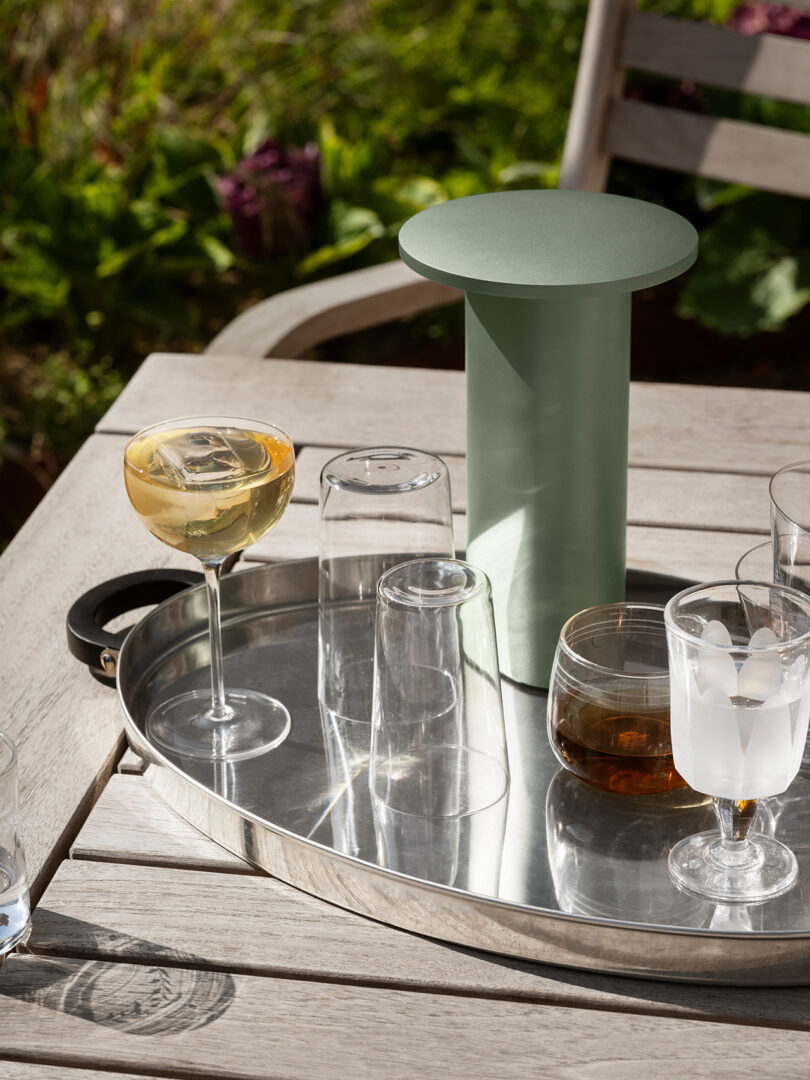 A green Solae lamp on a wooden outdoor table next to cocktails and greenery.