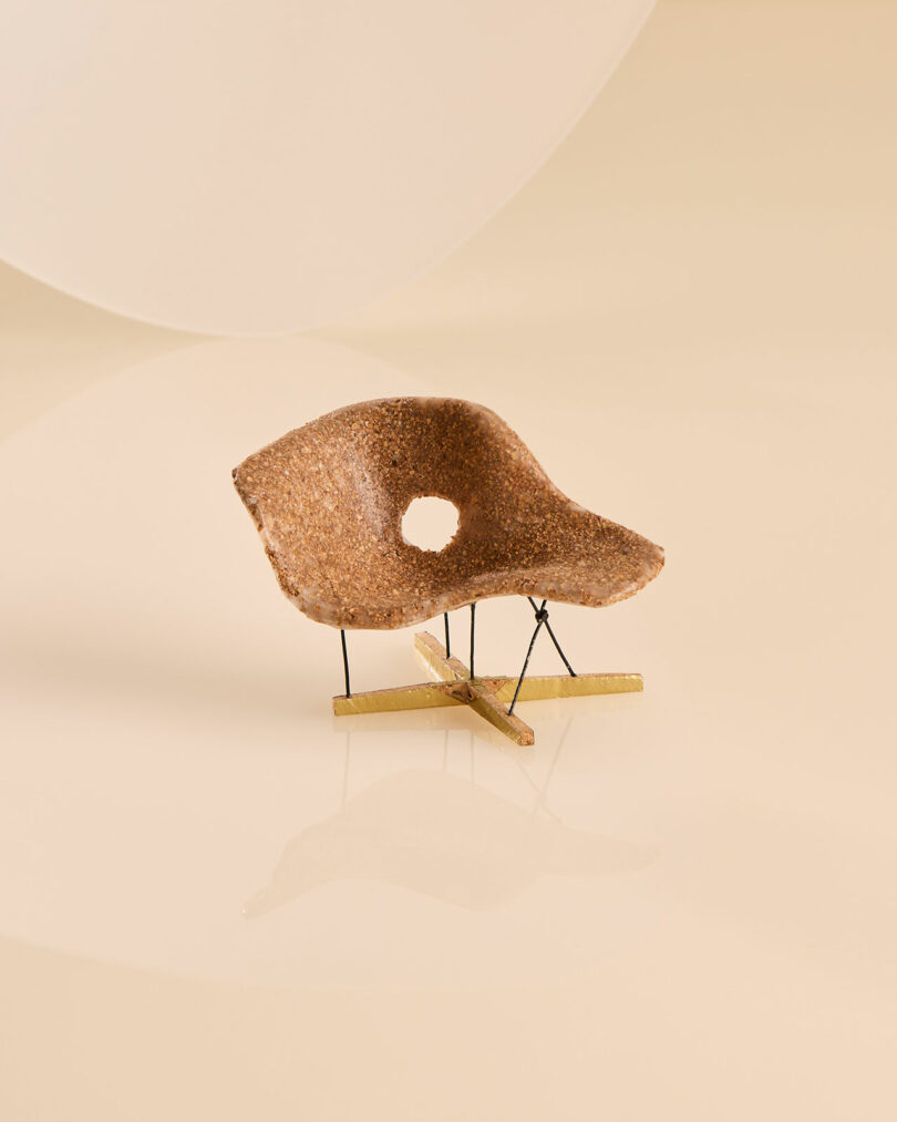 An abstract miniature chair sculpture with a hole in the center and thin wire legs.