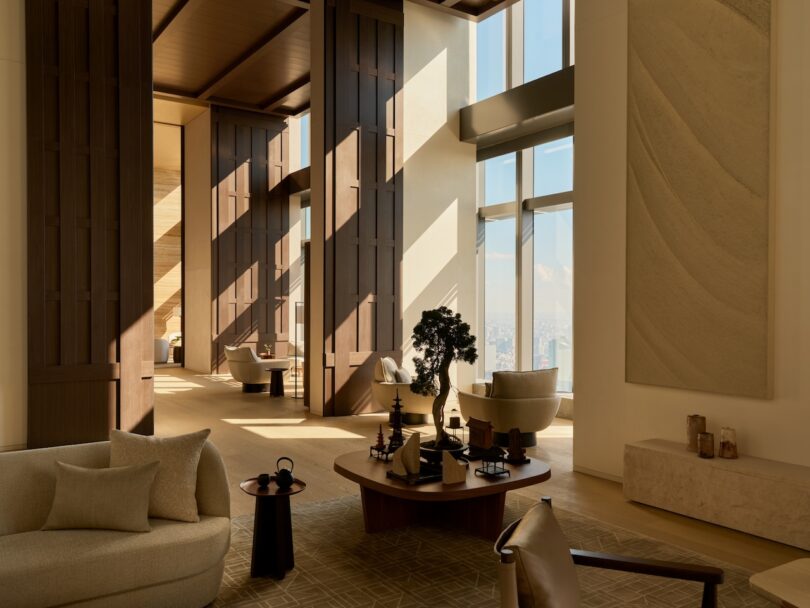 A spacious modern living room with beige sofas, high ceilings, and sunlight streaming through large windows.