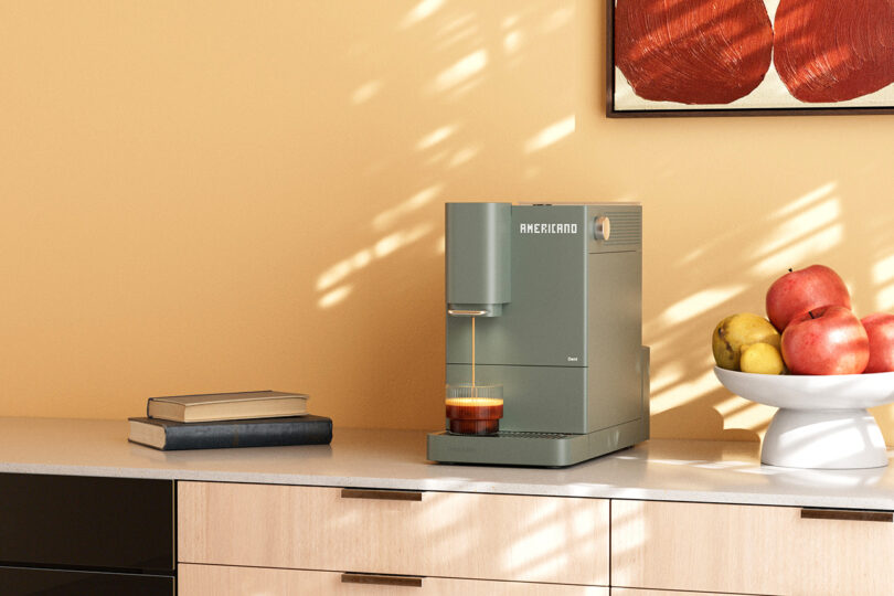 A wide shot of a coffee machine on a counter next to books and fruit, suggesting a quiet home routine.