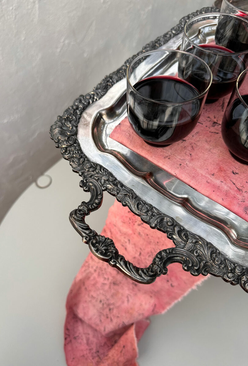 A close-up of an antique-style silver tray holding glasses of red wine against a soft pink fabric background.