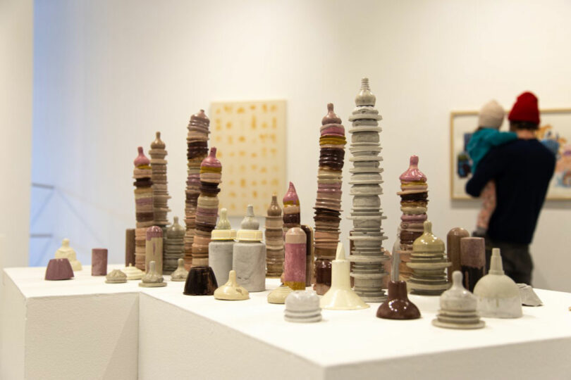 Ceramic sculptures on a pedestal in a gallery with a person holding a child in the background.