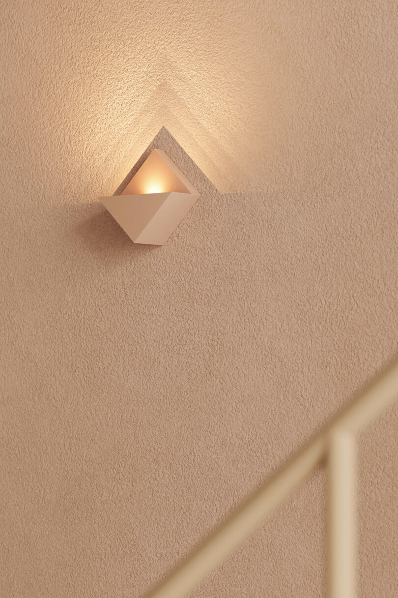 Modern wall sconce casting warm geometric light onto a textured beige wall near a staircase.