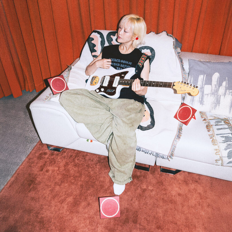 A person playing electric guitar on a couch surrounded by small red modular speakers.