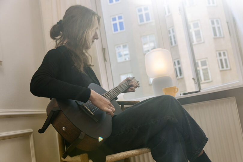 A person playing an acoustic guitar in a sunlit room near a window with a cup of coffee nearby.