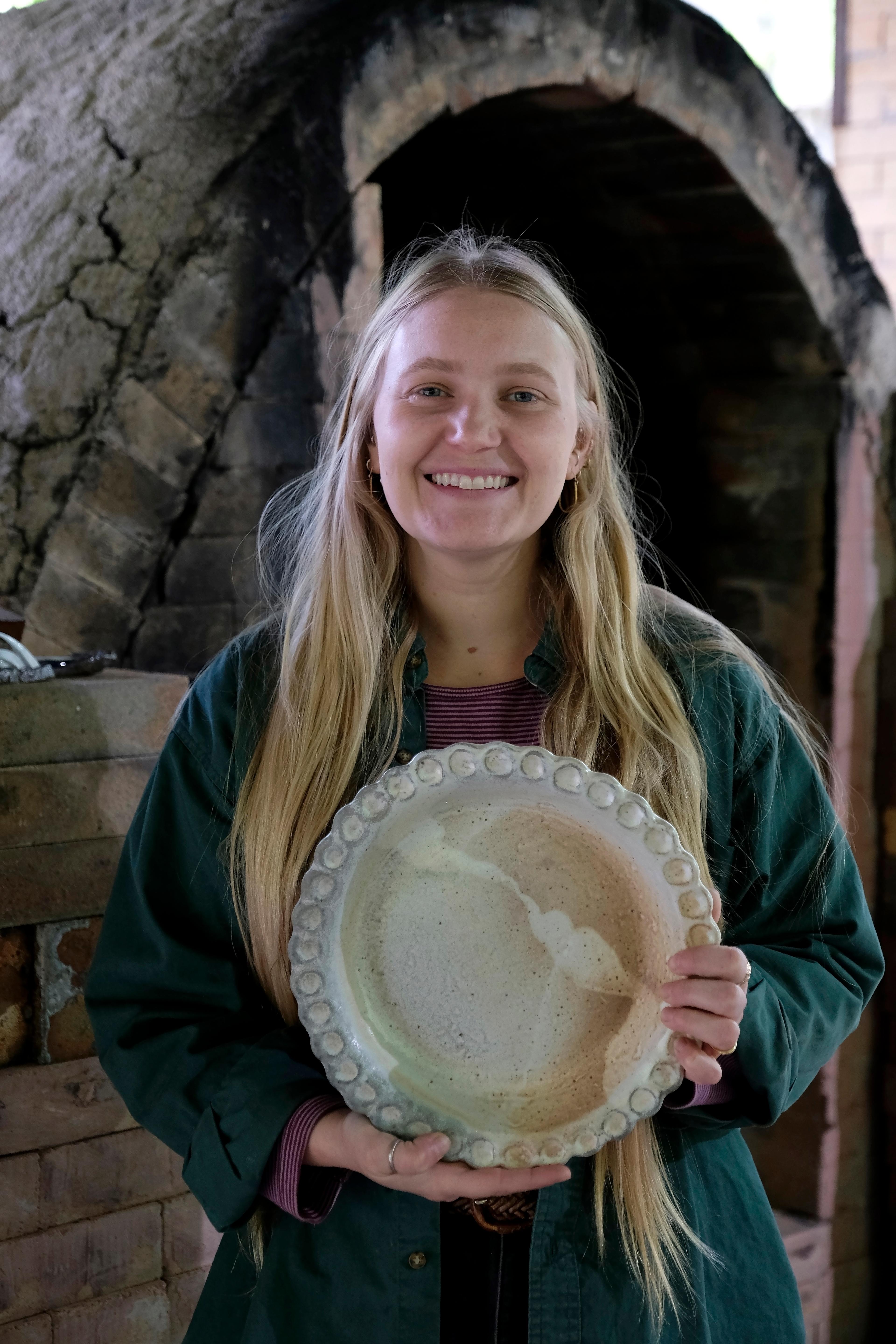 An artist holding a large, finished wood-fired ceramic pie plate with visible flame-flashing marks.