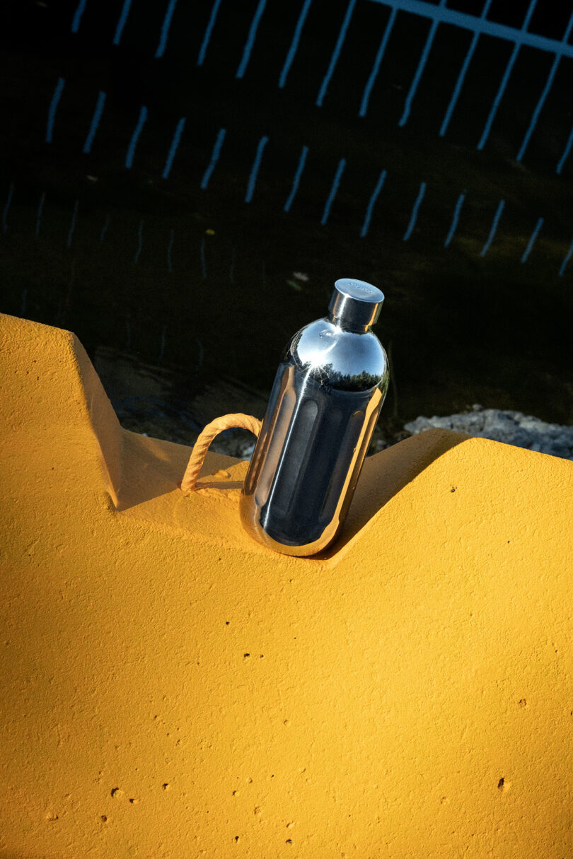 A reflective Aarke bottle on a bright yellow surface with a nautical rope and water in the background.