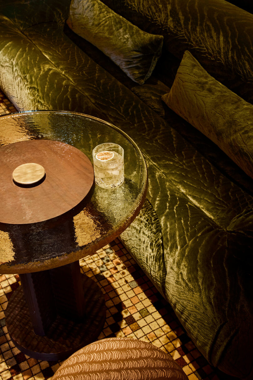 A cocktail glass on a glass table next to a green velvet sofa and patterned floor tiles.