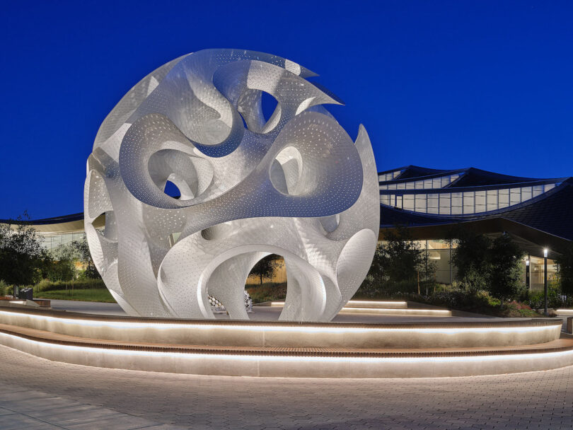 The metallic spherical sculpture illuminated from within at night on an outdoor plaza.