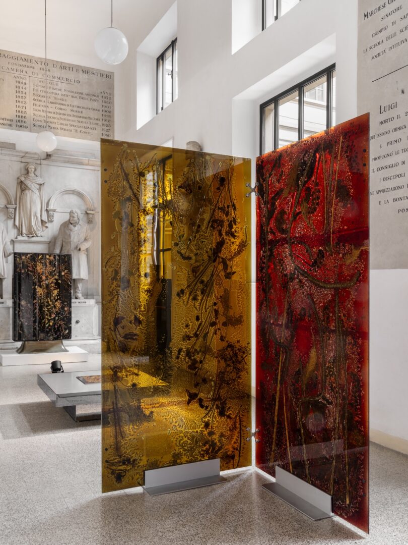 Two tall, vertical glass panels with embedded botanical silhouettes standing in a sunlit gallery.