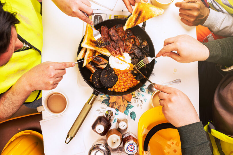Construction workers in high-visibility vests eating a breakfast of bacon and beans out of a Staub skillet.