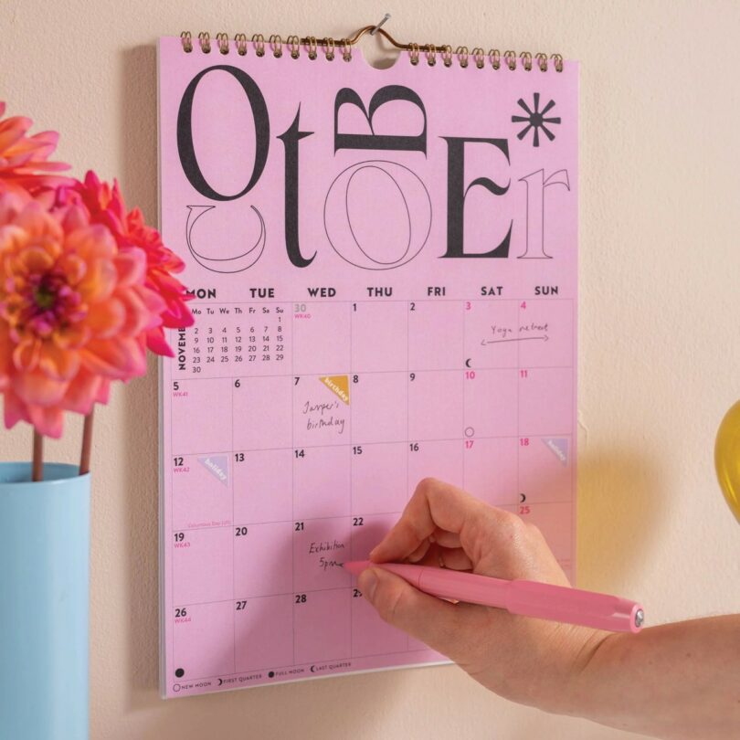 A close-up of a person writing a reminder on a pink 2026 wall calendar hanging next to a vase.