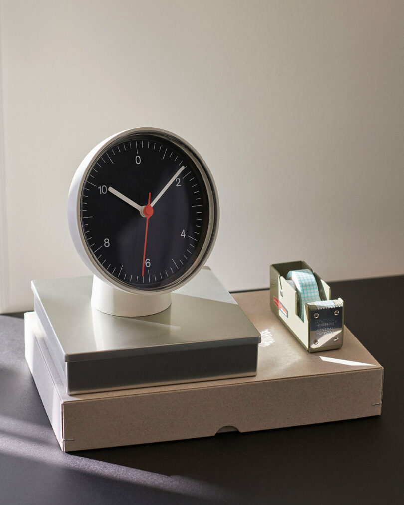 A round white analog table clock sitting on boxes next to a tape dispenser.