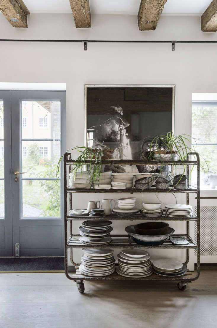 Eclectic ceramic collection stored on antique wooden shelves with wheels in a bright kitchen.