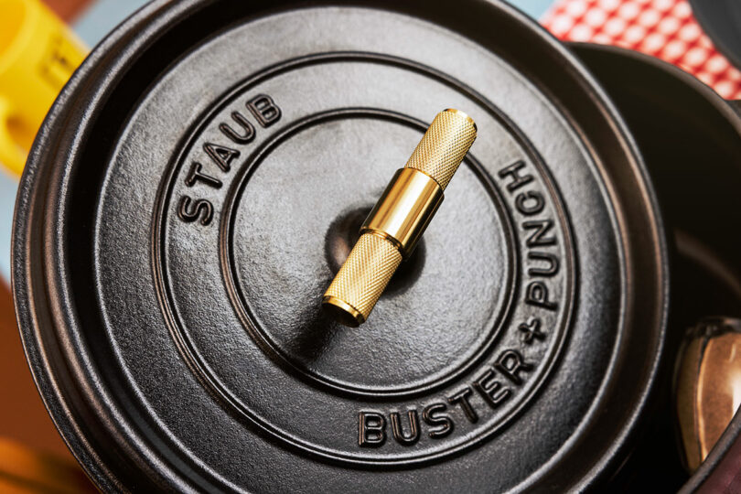 Close-up of a black Staub pot lid featuring a gold knurled handle and the embossed Buster + Punch logo.