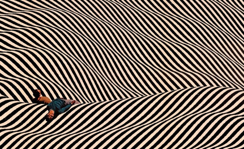 A person lying on a floor with high-contrast black and white wavy stripes creating a surreal optical illusion.
